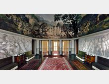 Salone interno di un palazzo storico con grandi affreschi sul soffitto e sulle pareti superiori e bassorilievi bianchi lungo le pareti laterali. Al centro ci sono tappeti rossi, un lampadario a candele e porte vetrate che si aprono verso un corridoio illuminato.