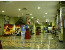 Atrio interno di un cinema con pavimento in marmo a mosaico, soffitto a pannelli con luci incassate e grandi poster di film su supporti espositivi. In fondo si vedono le indicazioni delle sale 2 e 3 e, sul lato destro, una fila di colonnine e corrimano metallici lungo le pareti.