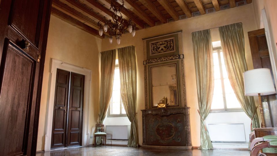 Interno di una stanza elegante con soffitto a cassettoni in legno, pavimento in parquet lucido e pareti tinte di giallo tenue. La stanza presenta due grandi finestre con tende verdi trasparenti, un camino decorato con specchio sovrastante e intagli floreali, oltre a una doppia porta in legno scuro.