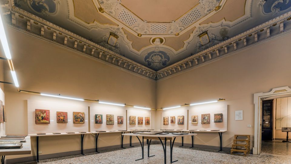 Sala espositiva in un edificio storico con soffitto alto decorato da stucchi e affreschi e pavimento in terrazzo. Lungo le pareti e su tavoli centrali sono esposte piccole opere e pannelli, illuminate da luci lineari a parete.