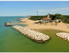 Veduta aerea di una costa sabbiosa con mare calmo e una serie di frangiflutti in pietra che proteggono piccole insenature. Sulla riva si trova un edificio con tetto in tegole e una torre faro bianca, circondato da vegetazione e dune.