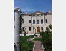 Cortile con prato e vialetto in mattoni che conduce a una fontana bassa davanti a un edificio in stile classico con frontone, alte finestre e portone centrale ad arco. In primo piano si vede una colonna in pietra e siepi curate ai lati del percorso.