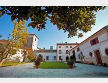 Cortile interno di una villa storica con prato centrale, ghiaia e vasi di terracotta, circondato da edifici intonacati con finestre e persiane rosse. Sullo sfondo si vedono una torre in pietra e tetti in coppi, con rami di un grande albero che incorniciano la scena dall’alto.