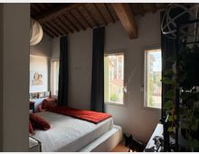 Camera da letto luminosa con letto matrimoniale, biancheria bianca e coperta rossa, pareti chiare e soffitto in legno con travi a vista. Due finestre con tende scure affacciano su edifici vicini, con una parete decorata da piante e una sfera luminosa sospesa.