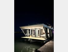 Casa galleggiante moderna ormeggiata a un pontile di legno di notte, con ampie vetrate e veranda illuminata affacciata sull’acqua. Interni visibili con tende bianche e arredi, sullo sfondo cielo scuro e luci lontane sulla riva.