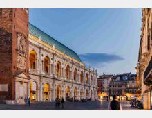 Piazza urbana al crepuscolo con un grande edificio storico in stile palladiano, caratterizzato da due ordini di arcate illuminate e un tetto verde. Ai lati si vedono facciate di palazzi cittadini e un’ampia pavimentazione aperta verso il centro della piazza.
