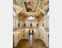Interno di un salone storico a doppia altezza con balconata perimetrale e balaustra in pietra, pareti ornate da affreschi e finte architetture. Volta e soffitto a cassettoni dipinti con scene figurative, grandi finestre ad arco e pavimento in terrazzo al centro dello spazio.