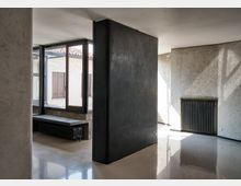 Interno di un edificio moderno con pareti in intonaco chiaro e un volume centrale scuro che divide lo spazio. Ampie finestre con telai scuri affacciano su un cortile, mentre radiatori a vista e pavimento lucido riflettente completano l’ambiente.