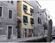 Vista di un canale urbano fiancheggiato da edifici in mattoni e intonaco, con un palazzo stretto al centro dalle finestre con persiane verdi e fioriere. Lungo la riva corre una banchina con paletti e un grande palo di legno vicino all’acqua.
