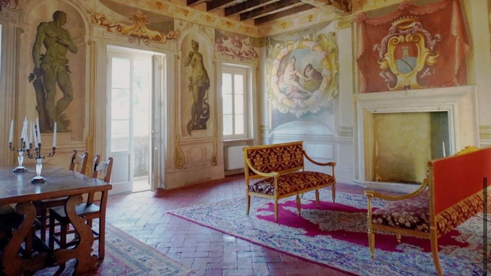 Salone storico con pareti affrescate e decorazioni barocche, soffitto a travi in legno e lampadario centrale. Nella stanza si vedono un grande camino, porte-finestre luminose, pavimento in cotto e arredi d’epoca con tappeti.