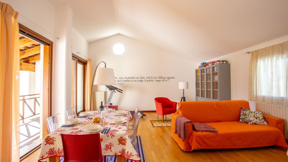 Soggiorno open space mansardato con pavimento in legno, tavolo da pranzo apparecchiato vicino a grandi porte finestre che danno su un balcone e un’area relax con divano arancione e poltrona rossa. Sul fondo si vedono una libreria/vetrina grigia, lampade da terra e una finestra con tende leggere.