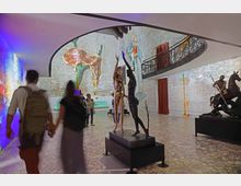 Interno di una galleria d’arte con pareti in mattoni bianchi e soffitto ad arco, con balconata superiore in ferro battuto. Nella sala sono esposte sculture su piedistalli e installazioni sospese, tra cui un grande orologio molle, con tende rosse e pavimento in pietra.