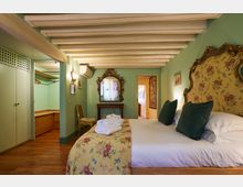 Camera da letto in stile classico con pareti verde chiaro e soffitto con travi a vista bianche, dominata da un grande letto con testiera decorata e copriletto floreale. Nell’ambiente si vedono armadio e cabina guardaroba aperta, specchiera ornamentale su consolle e pavimento in legno.