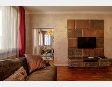 Soggiorno con pavimento in legno, grande finestra con tende bianche e drappi rossi e divano in primo piano. Sulla parete di fronte c’è un mobile TV con pannelli decorativi in tonalità rame e, attraverso un’apertura, si vede una sala da pranzo con lampadario e tavolo apparecchiato.