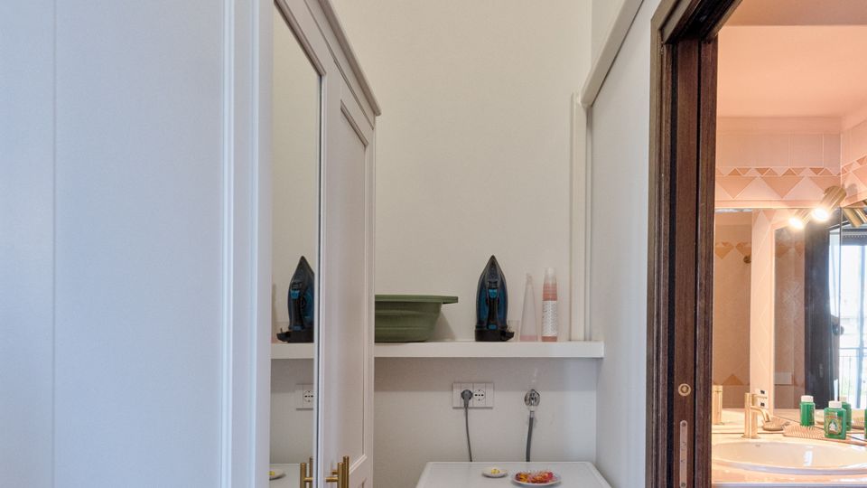 Piccolo ripostiglio/lavanderia con pavimento in legno, armadi bianchi a tutta altezza e lavatrice sotto una mensola con ripiani. A destra si vede l’apertura verso un bagno con lavabo e specchio illuminato.