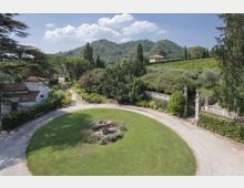 Vista dall’alto di un giardino di villa con prato circolare e fontana centrale, circondato da un vialetto in ghiaia. Sullo sfondo si vedono edifici con tetti in coppi, cipressi e colline verdi con vigneti e alberi sotto un cielo parzialmente nuvoloso.