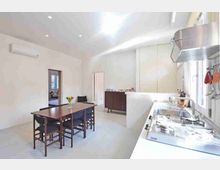 Cucina-soggiorno moderna e luminosa con pareti bianche, tavolo da pranzo in legno con sedie scure e armadi a tutta altezza. In primo piano si vede il piano cottura e il lavello in acciaio sotto una finestra, con cappa metallica e illuminazione a incasso sul soffitto.