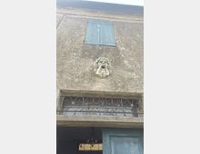 Facciata di un edificio in intonaco invecchiato con una piccola finestra chiusa da persiane azzurre e, sotto, una decorazione in pietra a forma di mascherone. Più in basso si vede un balcone con ringhiera in ferro battuto e parte di un portone in legno con lampadario all’interno.