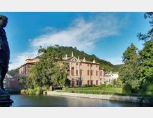 Villa storica con facciata rosa e statue sul tetto affacciata su un canale, circondata da giardini curati con siepi e alberi. Sullo sfondo si vedono colline boscose e un cielo sereno.
