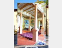 Portico interno di una villa storica con colonne in pietra e soffitto a travi dipinte, pavimento in cotto e tappeti. Sul fondo si vedono pareti affrescate con grandi medaglioni decorativi e un passaggio verso una sala arredata.