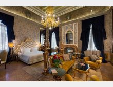 Interno di una camera da letto in stile classico con letto matrimoniale dal grande testiero imbottito, lampadario in vetro dorato e pareti decorate con affreschi e motivi damascati. Grandi finestre con tende scure, camino con specchiera e zona salotto con divani e tavolini completano l’ambiente.