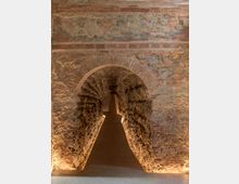 Corridoio sotterraneo in pietra con passaggio ad arco in mattoni, pareti irregolari in muratura e illuminazione a pavimento lungo i lati. Il tunnel stretto conduce verso un’apertura interna sul fondo.