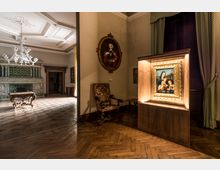 Interno di una sala museale in un palazzo storico, con pavimento in parquet a spina di pesce, soffitti decorati e lampadari in vetro. In primo piano una teca illuminata con un dipinto incorniciato, accanto a una poltrona antica, mentre sullo sfondo si vede un grande camino in maiolica verde e un tavolo intagliato.