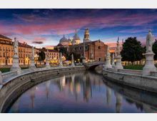 Canale urbano curvo al tramonto con parapetti in pietra e statue lungo le rive, attraversato da un piccolo ponte. Sullo sfondo si vede una grande basilica con cupole e campanile, circondata da edifici storici e alberi, con riflessi sull’acqua.