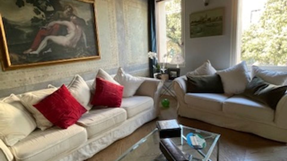 Soggiorno luminoso con due grandi divani chiari e cuscini decorativi, tavolino in vetro e cornice a parete. Finestre ampie con vista sul verde, pavimento in legno e soffitto con travi a vista.