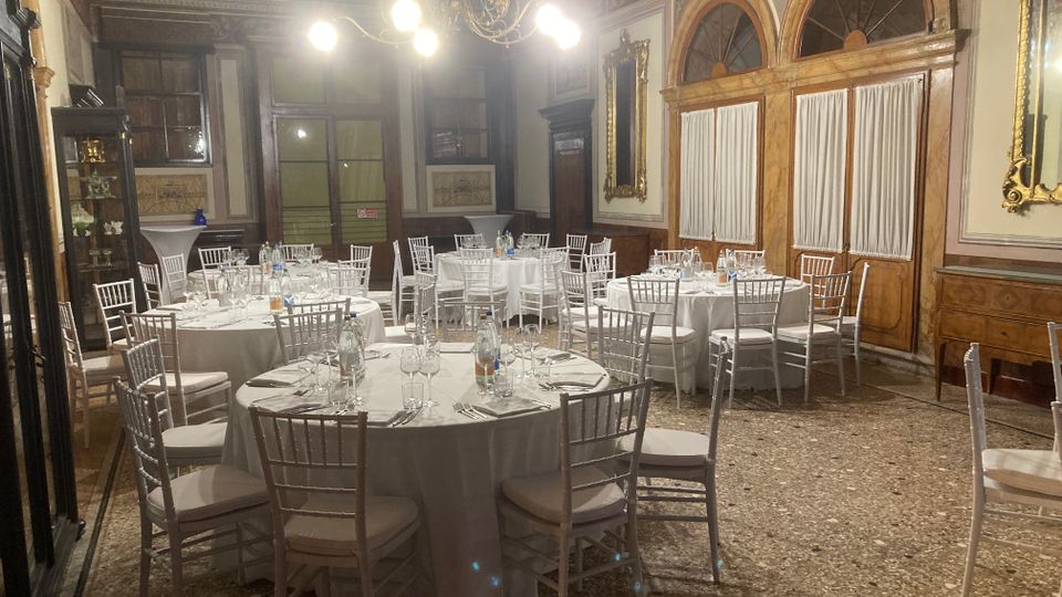 Sala per banchetti in stile classico con tavoli rotondi apparecchiati con tovaglie bianche, bicchieri e posate, circondati da sedie bianche. Pareti con boiserie in legno, grandi porte ad arco con tendaggi, specchi dorati e lampadario centrale sopra un pavimento in terrazzo.