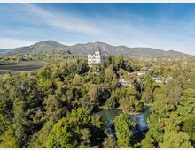 Veduta aerea di una villa storica bianca con torre centrale, immersa in un grande parco alberato con laghetto. Sullo sfondo si vedono campi e un’area collinare/montuosa sotto un cielo sereno.