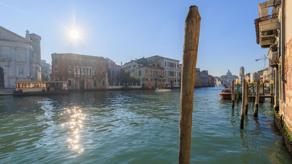 Veduta di un ampio canale urbano con acqua verde e riflessi del sole, fiancheggiato da palazzi storici e una chiesa sulla sinistra. In primo piano si vede un palo d’ormeggio e, sulla destra, una fila di pali e piccole imbarcazioni lungo la riva.