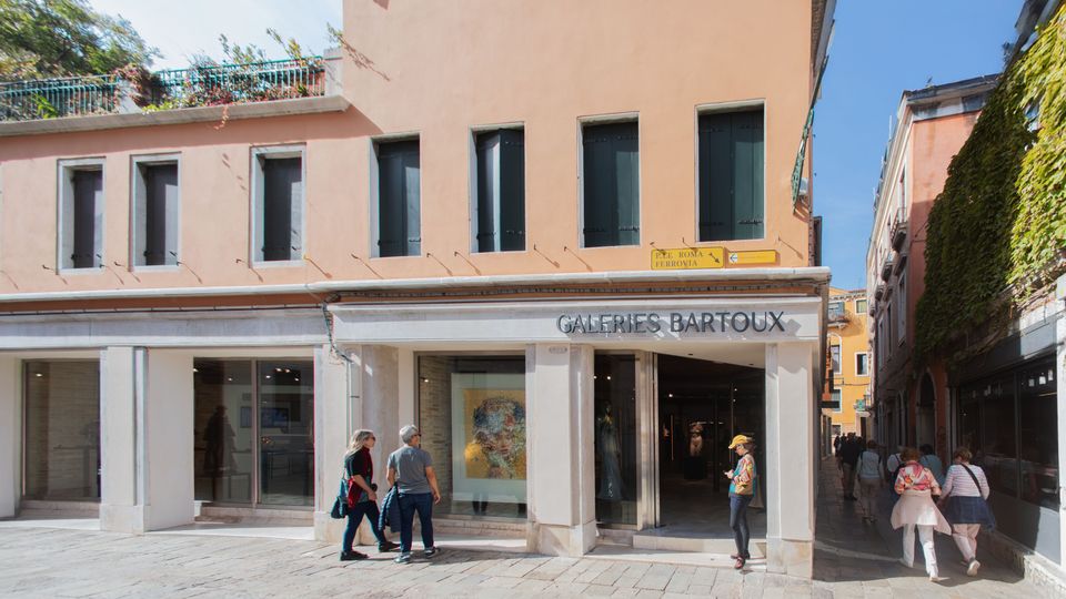 Facciata di un edificio color pesca con portico e grandi vetrine d’ingresso della galleria d’arte “Galeries Bartoux”, su una piazza pavimentata in pietra. A destra si apre una stretta via tra edifici, con un muro ricoperto di edera e segnaletica stradale sopra l’angolo dell’edificio.