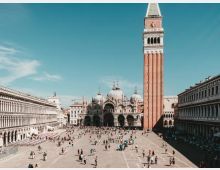 Ampia piazza lastricata circondata da lunghi palazzi porticati, con al centro la Basilica di San Marco dalle cupole ornate e, a destra, l’alto campanile in mattoni. Sullo sfondo si vedono edifici storici e un cielo sereno sopra lo spazio aperto della piazza.