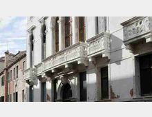 Facciata di un edificio storico in pietra chiara con alte finestre ad arco e balconata decorata a rilievo, sopra un ingresso centrale ad arco. La strada urbana è affiancata da altri edifici più bassi con tetti in tegole sul lato sinistro.