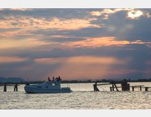 Imbarcazione bianca su una laguna con palificate e strutture da pesca in legno che emergono dall’acqua. Sullo sfondo, orizzonte piatto con colline lontane e cielo ampio e nuvoloso illuminato dalla luce del tramonto.