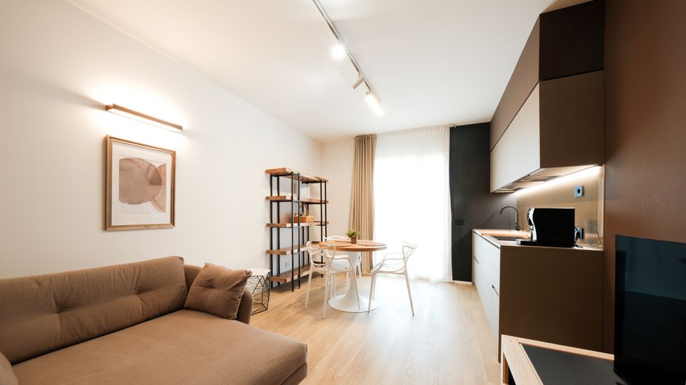 Open space moderno con soggiorno e cucina lineare a vista: divano beige, pavimento in legno chiaro e tavolo rotondo con sedie vicino a una grande finestra con tende. Pensili scuri e bianchi illuminati da luci a binario e una scaffalatura metallica completano l’ambiente.