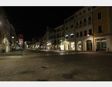 Piazza urbana lastricata in pietra di notte, fiancheggiata da edifici storici con portici e vetrine illuminate. Sullo sfondo si vede una torre con orologio e luci rosse, con lampioni che illuminano la strada ampia e quasi vuota.