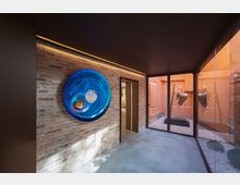 Interno di uno spazio espositivo moderno con parete in mattoni a vista e grande installazione circolare in vetro blu, pavimento in cemento e soffitto scuro. Sulla destra, una parete vetrata affaccia su un piccolo cortile con ghiaia e un’opera scultorea metallica su base nera.