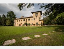 Ampio prato con un percorso di pietre in primo piano davanti a una grande villa storica in pietra, con finestre ad arco, torretta e balconi. L’edificio è circondato da alti alberi e giardini, sotto un cielo parzialmente nuvoloso.