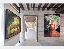 Interno di una galleria d’arte con pareti in mattoni a vista, pavimento in cemento e soffitto con travi di legno e tubazioni industriali. Nel corridoio centrale si vedono grandi quadri appesi alle pareti e un’apertura che conduce a un’altra sala espositiva illuminata da faretti.