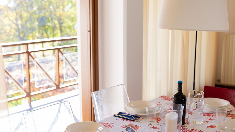 Sala da pranzo luminosa con tavolo apparecchiato, tovaglia a motivi marini, piatti bianchi e bicchieri, con lampada da tavolo e bottiglie al centro. Accanto a una grande porta-finestra che si apre su un balcone con ringhiera in legno e tettoia, affacciato sul verde esterno.