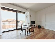 Soggiorno moderno e luminoso con pavimento in legno, pareti bianche a boiserie e zona pranzo con tavolo rotondo e sedie. Ampia portafinestra scorrevole con tende che apre su un balcone con ringhiera e vista sui tetti, con lampada a sospensione e piccolo mobile con TV.