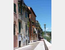 Strada pedonale lungo un lungofiume con muretto e lampione, fiancheggiata da case a più piani con facciate colorate e intonaco consumato, balconi e persiane. Sullo sfondo si vede il paesaggio collinare sotto un cielo sereno.