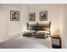 Camera da letto minimalista con letto matrimoniale, testiera in metallo scuro e biancheria grigia. Pareti bianche con tre fotografie incorniciate sopra il letto, comodino nero con lampada da lettura e ciotola in legno.