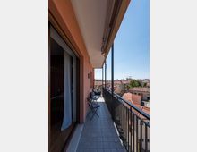 Balcone lungo e stretto di un appartamento con pavimento piastrellato, ringhiera metallica nera e tenda da sole superiore. Sono presenti un piccolo tavolo e sedie pieghevoli e la vista si apre sui tetti in tegole rosse e sugli edifici della città sotto un cielo sereno.