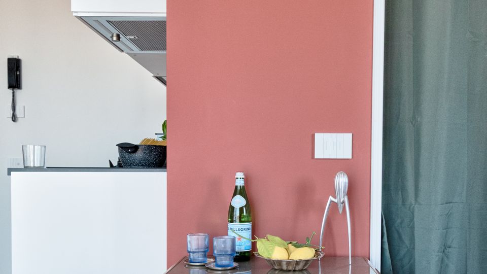 Interno domestico con parete color terracotta e pavimento in legno, con un piccolo mobile in legno a due cassetti appoggiato al muro. A sinistra si intravede una cucina moderna bianca con cappa, mentre a destra una porta con tenda verde scuro.