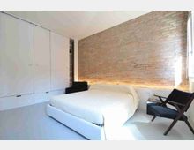 Camera da letto moderna e minimale con letto matrimoniale bianco, grande parete in mattoni a vista con illuminazione radente e armadiatura a tutta parete in pannelli bianchi. Ampia finestra laterale che illumina la stanza, con due poltroncine scure e un piccolo tavolino vicino al letto.