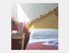 Camera da letto con letto matrimoniale con testiera decorativa e copriletto chiaro; accanto c’è un comodino in legno vicino a una finestra luminosa con tende chiare.