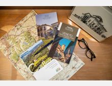 Piano in legno con mappe cartacee del Veneto e materiali turistici, tra cui guide e brochure, disposti a ventaglio. Sono visibili anche una penna, un paio di occhiali e una stampa incorniciata con disegno architettonico.